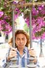Продуманий модний чоловік з квіткою в довгому волоссі шліфується біля паркану в парку в місті і дивиться на камеру — стокове фото