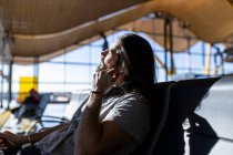 El tipo en el sombrero en el aeropuerto en la sala de espera sentado esperando su vuelo, se pone auriculares inalámbricos para escuchar música - foto de stock
