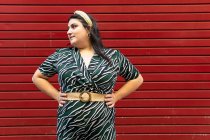Portrait of cheerful young curvy brunette in stylish striped outfit and headband while looking away against red wall — Stock Photo
