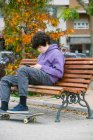 Vista lateral del adolescente masculino concentrado sentado en un banco de madera con monopatín mientras navega por Internet en el teléfono celular en el parque en la calle - foto de stock