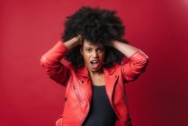 Freche Afroamerikanerin schreit und berührt Haare, während sie in die Kamera auf rotem Hintergrund im Studio schaut — Stockfoto