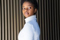 Side view of unemotional African American female in trendy sweater looking away against striped building wall on street — Stock Photo