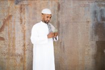 Fröhlicher muslimischer Mann in traditioneller Kleidung, der lächelt und in der Nähe einer schäbigen Mauer auf der Straße sein Handy schmökert — Stockfoto