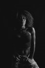 Кудрявая афроамериканка в модном топе и джинсах, стоящих с рукой на талии на черном фоне в студии — стоковое фото