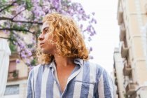 From below of dreamy curious male with long wavy hair standing in street in summer with flower tree — Stock Photo