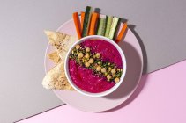 Top view of appetizing beetroot hummus garnished with chickpea served on two colored background with bread and fresh carrot and cucumber sticks — Stock Photo