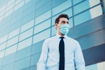 Снизу предприниматель-мужчина в формальной одежде с медицинской маской, смотрящий в город во время пандемии коронавируса — стоковое фото