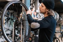Visão lateral do jovem mestre masculino examinando pneu na roda de bicicleta enquanto trabalhava na oficina de serviço de reparo profissional — Fotografia de Stock