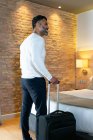 Вид сзади низкий угол положительный этнический мужчина путешественник с багажом стоя возле кровати в номере отеля — стоковое фото