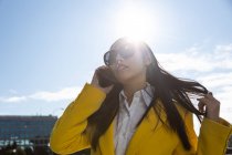 Sorridente donna d'affari asiatica con cappotto giallo e smart phone che cammina per strada con edificio sullo sfondo — Foto stock