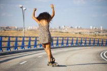 Mujer joven y afro patinaje tabla larga por un puente vacío al atardecer, vista trasera - foto de stock