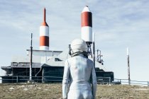 Mann im Raumanzug steht an sonnigem Tag auf felsigem Boden gegen Metallzaun und gestreifte raketenförmige Antennen — Stockfoto