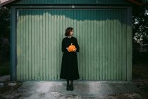 Female in black dress and with Halloween pumpkin lantern standing against wooden wall of house and looking away — Stock Photo