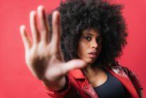Verängstigte Afroamerikanerin mit lockigem Haar zeigt Stopp-Geste auf rotem Hintergrund im Studio und blickt in die Kamera — Stockfoto