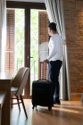 Full body low angle side view of adult man with luggage opening curtain hanging on balcony door in hotel room — Stock Photo