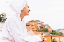 Ottimista giovane donna in accappatoio e asciugamano sorridente e distogliendo lo sguardo mentre si rilassa sul balcone durante la routine di cura della pelle nel fine settimana — Foto stock
