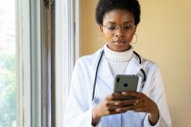 Серьёзная молодая афроамериканка-врач в белом халате со стетоскопом проверяет информацию на мобильном телефоне, стоя у окна клиники — стоковое фото