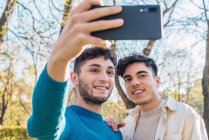 Alegre pareja de hombres homosexuales abrazando y tomando auto disparo en el teléfono móvil en el parque - foto de stock