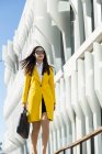 Азійська бізнес-жінка з жовтим пальто і розумний телефон ходити по вулиці з будівлею на задньому плані — стокове фото