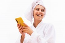 Mujer joven en albornoz y toalla sonriendo y navegando por el teléfono móvil mientras descansa en el balcón después de la ducha - foto de stock