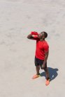 Low angle of thirsty African American athletic male drinking fresh water from plastic bottle during training on sunny day in city — Stock Photo