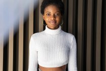 Stilvolle junge schwarze Frau im Crop-Pullover blickt in die Kamera, während sie in der Nähe einer gestreiften Hauswand in der Stadt steht — Stockfoto
