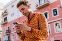 Снизу вид на молодого латиноамериканца с африканскими волосами в стильном красочном наряде, просматривающего мобильный телефон, стоя на перилах возле городского здания под солнечным светом — стоковое фото