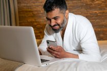 Crop sonriente hombre adulto en blanco albornoz netbook navegación y teléfono móvil mientras está acostado en la cama blanda en la habitación de hotel - foto de stock