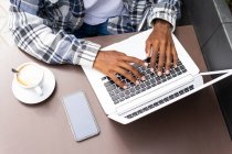 De cima cortado irreconhecível Africano americano masculino freelancer navegação e trabalhar remotamente no laptop no café ao ar livre, enquanto sentado à mesa com xícara de café — Fotografia de Stock