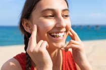 Sonriente hembra aplicando loción bronceadora en la cara en un día soleado en verano en la playa - foto de stock
