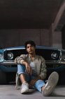Retrato de jovem latino em roupas casuais olhando com confiança para a câmera enquanto sentado inclinado em um carro vintage no estacionamento — Fotografia de Stock