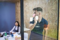 Gerente ejecutivo masculino escribiendo en nota adhesiva en la pared de vidrio durante la lluvia de ideas con compañeros de trabajo en la oficina - foto de stock