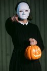 Spooky anonymous female in white Halloween mask and with glowing jack o lantern pointing at camera while standing in city — Stock Photo