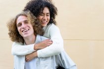 Fröhlicher Mann gibt seiner Freundin Huckepack-Fahrt, beide mit lockigem Haar und lautem Lachen — Stockfoto