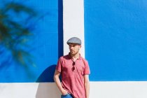 Задумчивый молодой бородатый мужчина в стильной повседневной одежде и кепке наслаждается летним днем возле голубой стены на городской улице — стоковое фото