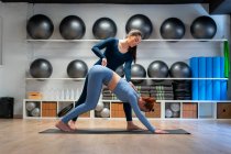 Longitud completa de hembra joven haciendo postura hacia abajo con la ayuda de entrenador durante la clase de yoga en el gimnasio - foto de stock