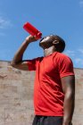Low angle of thirsty African American athletic male drinking fresh water from plastic bottle during training on sunny day in city — Stock Photo