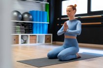 Voller Körper einer ruhigen, konzentrierten jungen Frau, die in Heldenpose mit namaste Händen sitzt, während sie Achtsamkeit und Meditation während eines Yoga-Kurses im Fitnessstudio übt — Stockfoto