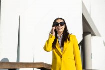 Lächelnde asiatische Geschäftsfrau mit gelbem Mantel und Smartphone, die auf der Straße läuft, mit einem Gebäude im Hintergrund — Stockfoto