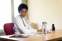 Giovane medico afroamericano in vestaglia bianca con stetoscopio seduto a tavola con laptop e lettura di cartelle cliniche mentre lavora in ambulatorio — Foto stock