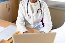З - над врожаю афроамериканська жінка - лікар надрукує звіт про ноутбук, працюючи за столом у сучасній клініці. — стокове фото