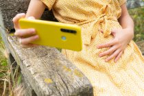 Обрезанный неузнаваемая беременная женщина в платье трогательный живот и сделать автопортрет на мобильном телефоне, сидя на скамейке в сельской местности летом — стоковое фото