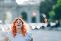 Positive charming female with ginger hair laughing with closed eyes in city street — Stock Photo