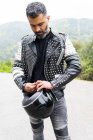 Serious brutal ethnic male biker in stylish leather jacket standing on asphalt road with helmet in hand near lush green trees growing in mountainous valley — Stock Photo