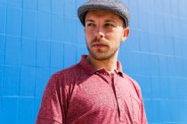 Thoughtful young bearded male in stylish casual wear and cap enjoying summer day near blue wall on urban street — Stock Photo