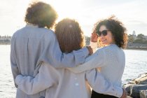 Back view of anonymous friends with curly hair standing close in hug against cityscape and sea in sunlight — Stock Photo