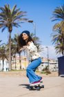 Corpo inteiro de fêmea feliz ativa em roupas casuais andando de skate na estrada ao longo da praia de areia e palmas altas durante o treinamento — Fotografia de Stock