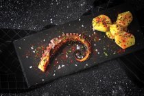 From above of fried octopus tentacle and pieces of potato served with spices on black board on table — Stock Photo
