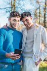 Delighted homosexual couple of males embracing and watching funny video on mobile phone while standing in park and having fun — Stock Photo