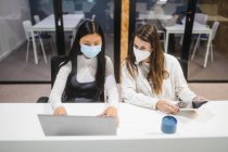 Multiracial female colleagues wearing masks sitting at table with laptop and discussing business project while working in coworking space — Stock Photo